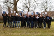 Personen stehen für ein Gruppenfoto auf einer Wiese und halten Schriftstücke in die Luft